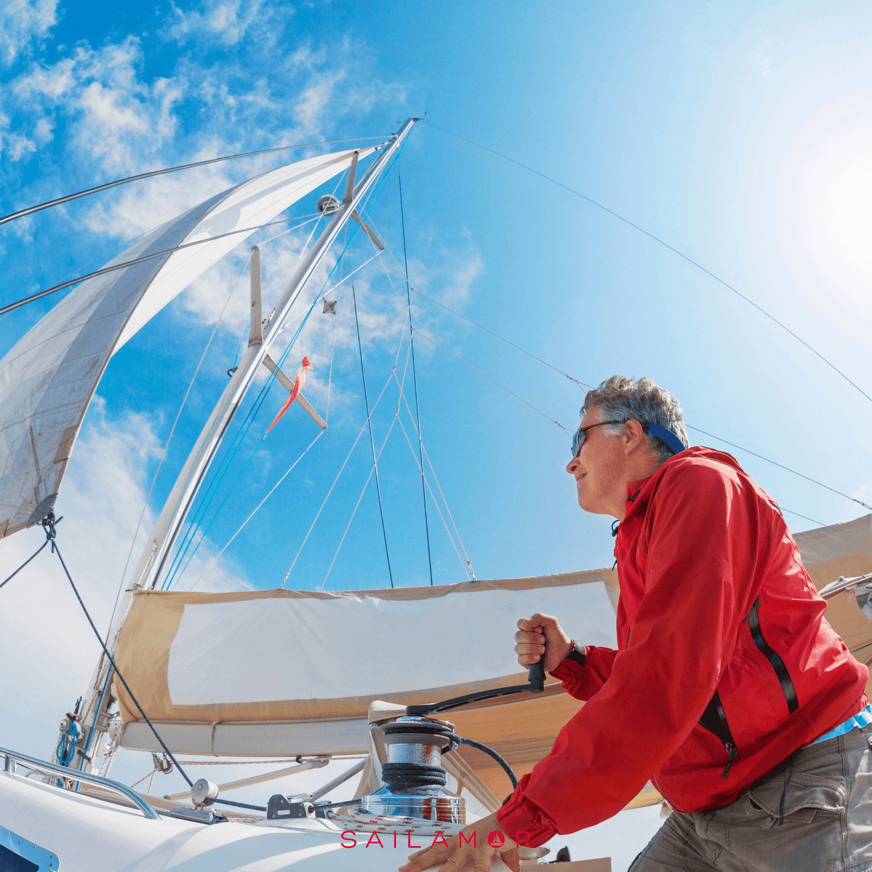 Man in a red jacket operating a winch on a sailboat under a clear blue sky.